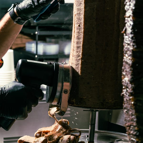 A chef in black gloves uses an electric knife to shave steaming shawarma meat from a vertical spit.