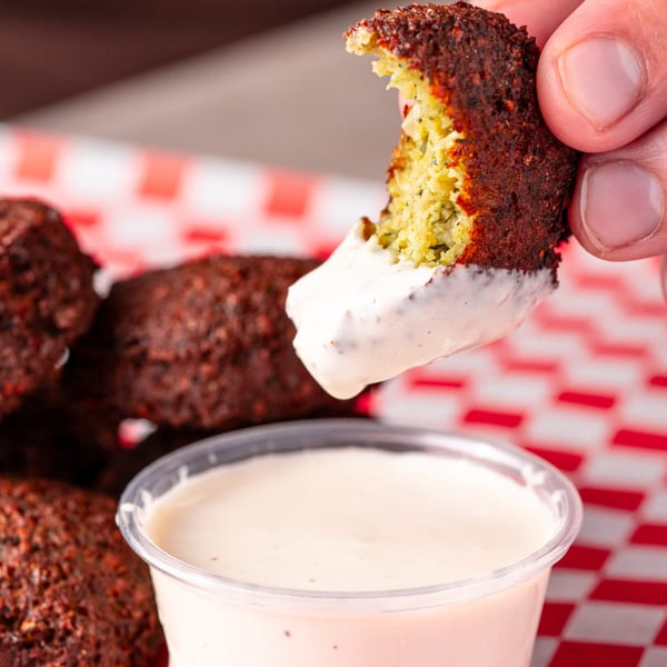 Hand dipping a Shawarma Shack falafel into a white sauce, with more falafel in a red checkered basket.