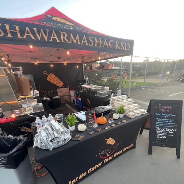 Shawarma Shack's outdoor catering setup with a shawarma machine, food, and menu board.