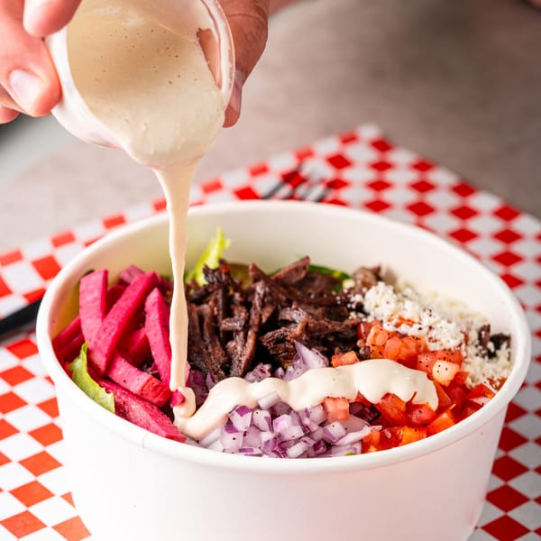 Creamy white sauce being poured over a Shawarma Shack bowl with meat, pickled turnips, and vegetables.
