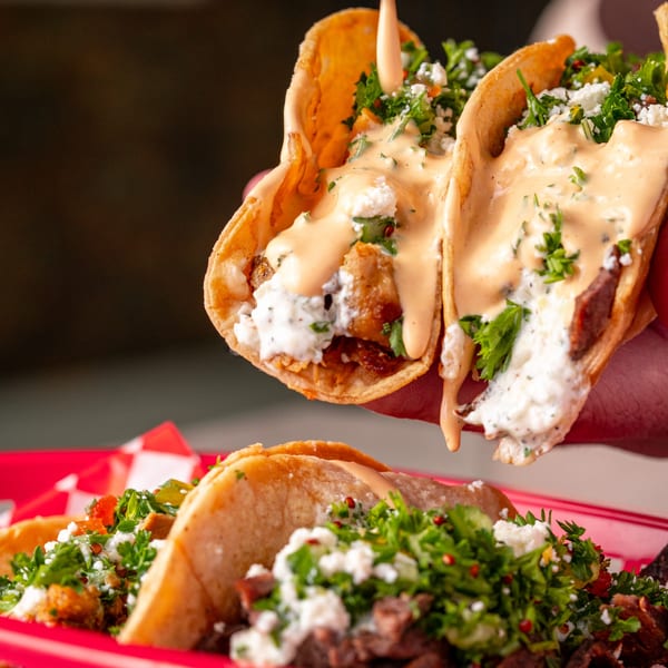 Hand holding two Shawarma Shack tacos with meat, feta, parsley, and sauce, with more tacos on a red plate.